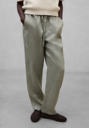 Person som har på seg løstsittende, lyse olivengrønne bukser med snor, kremfarget strikkegenser og mørkebrune, uformelle sko, stående med hendene i lommene.