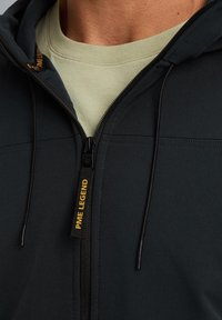 Donkergrijze zip-up hoodie met zwarte rits en gele "PME LEGEND" label. Beschikt over een textuur oppervlak en een vastgenaaide capuchon. Daaronder een lichtbeige t-shirt.