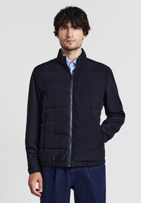 Homme aux cheveux bouclés foncés portant une veste noire matelassée zippée sur une chemise à col bleu clair sur fond uni.