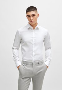 White long-sleeve shirt in smooth fabric with a pointed collar and button front, paired with light gray textured trousers.