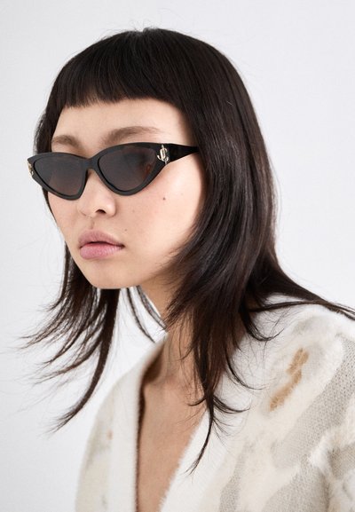 Lunettes de soleil noires en forme de chat avec des verres teintés et des accents en or en forme d'ancre sur les côtés, portées par un modèle aux cheveux foncés de longueur moyenne.
