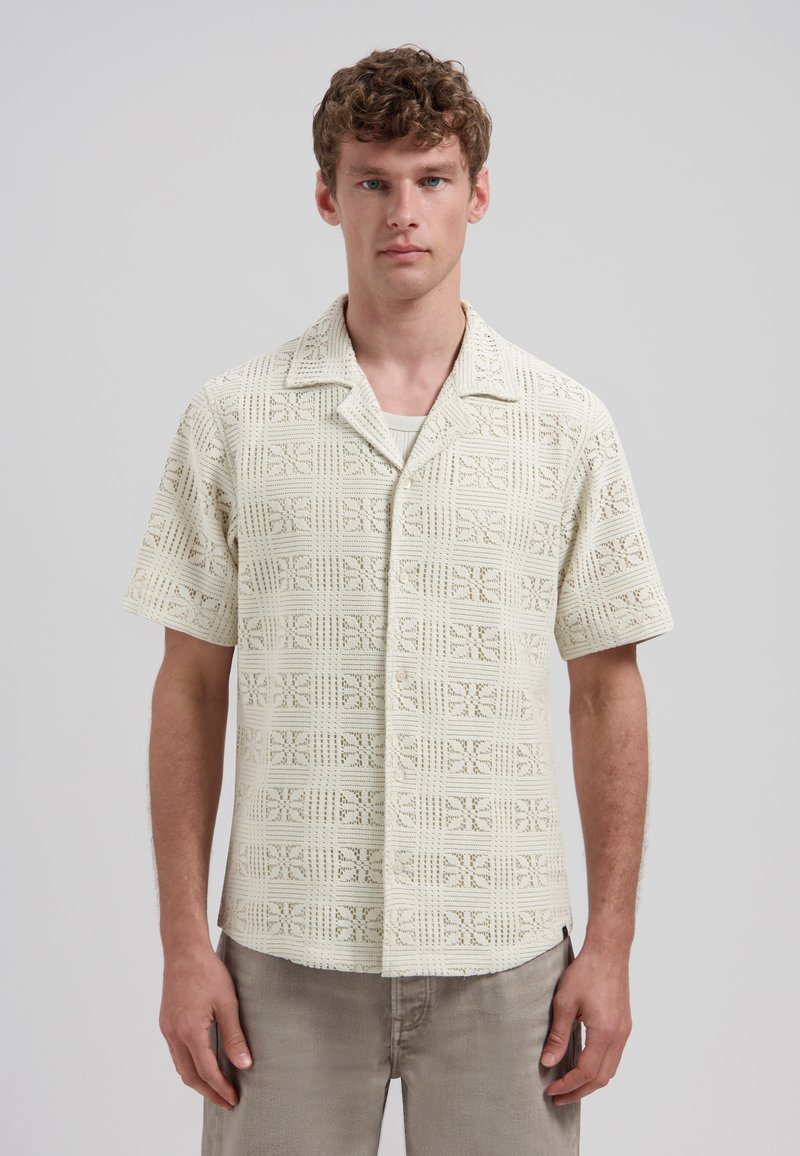 Jeune homme aux cheveux bouclés bruns portant une chemise à boutons en crochet crème à manches courtes et un pantalon gris clair, debout devant un fond uni.