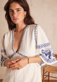 Abito bianco ricamato con motivi floreali blu su maniche e scollatura; tessuto morbido e strutturato con un design a scollo a V profondo. Accessori in gioielleria dorata.