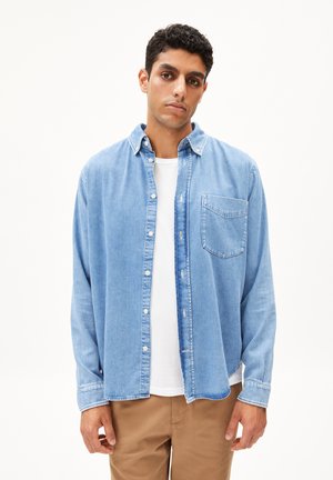 Light blue denim shirt with button closure, collar, and pocket on the chest, worn over a white t-shirt; paired with beige pants.