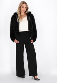 Schwarze Kunstpelz-Cropped-Jacke mit Kapuze und Reißverschluss, kombiniert mit hoch sitzenden, schwarzen weit geschnittenen Hosen und schwarzen, mit Nieten besetzten Absätzen.