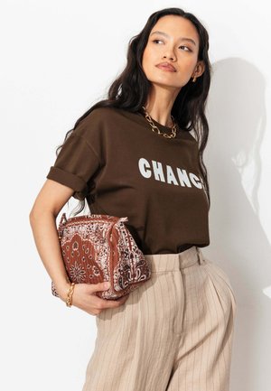 Femme aux longs cheveux foncés porte un T-shirt marron « CHANGE », un pantalon beige à fines rayures, un collier et un bracelet en chaîne dorée, tenant un sac marron à motif.