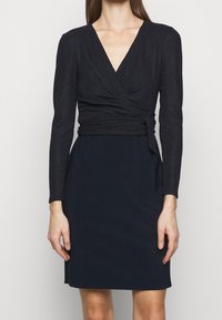 Černé šaty s výstřihem do V, dlouhými rukávy a vypasovaným korzetem se zavinovacím designem, přecházející do hladké sukně. Měkká textura.