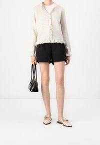 Beige stickad kavaj med volangkant och knappstängning, bära över en vit skjorta. Svarta shorts och beigea slip-on skor, håller en liten svart väska.