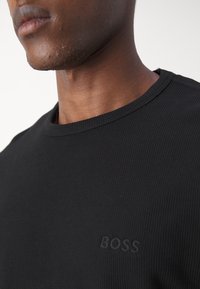 Gros plan d’une personne portant un t-shirt noir texturé à col rond avec le logo "BOSS" en relief sur la poitrine.