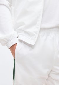 Άτομο που φοράει λευκό παντελόνι φόρμας και μπουφάν με το χέρι στην τσέπη, εμφανίζοντας λεπτομέρειες με ελαστικά τελειώματα στα μανίκια και στη μέση.