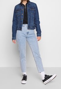 Tmavě modrá džínová bunda s knoflíkovými zapínáním, dvěma kapsami na hrudi a klasickým límcem. Kombinována se světlými modrými džínami s rovnými nohavicemi a černými teniskami.