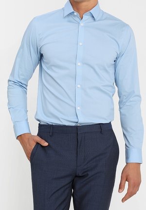 Man wearing a light blue button-up shirt tucked into dark navy dress pants, with one hand in his pocket and the other relaxed by his side.