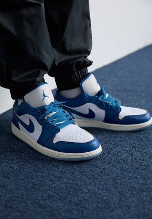 Personne portant des baskets Nike Air Jordan blanches et bleues avec des lacets bleus, debout sur un tapis bleu foncé, habillée d’un pantalon noir et de chaussettes blanches.