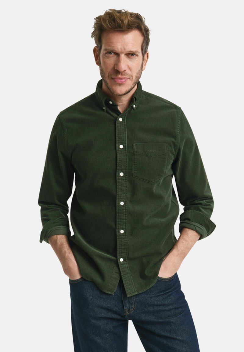 Camisa de botonadura en corduroy verde oscuro con un bolsillo frontal y botones blancos, combinada con jeans de mezclilla azul oscuro. Mangas largas enrolladas.