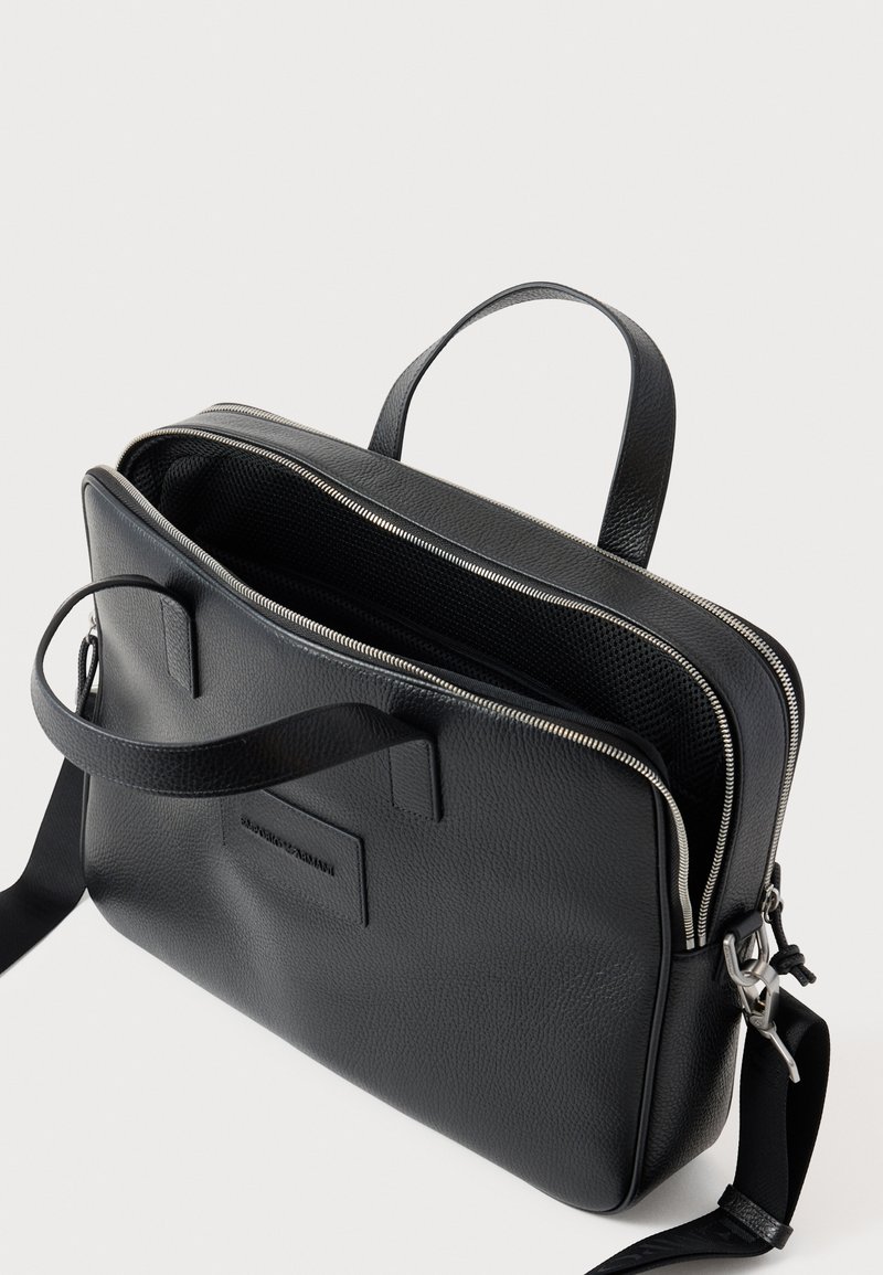 Borsa in pelle nera con texture liscia, dotata di chiusura a zip, due manici e tracolla regolabile. Presenza di un dettaglio con logo notevole.