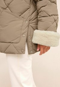 Chaqueta acolchada tipo puffer beige con un puño de felpa crema esponjoso, presenta un gran bolsillo lateral y cierres a presión, combinada con pantalones blancos.