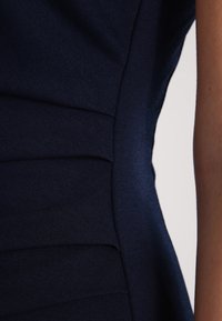 Robe bleu marine avec une coupe ajustée, un tissu texturé, des plis latéraux et une finition lisse le long de la couture pour un look structuré.