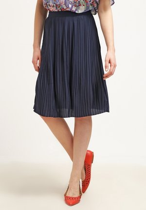 Femme portant une jupe plissée bleu marine associée à des ballerines rouges perforées, debout jambes croisées sur un fond clair.