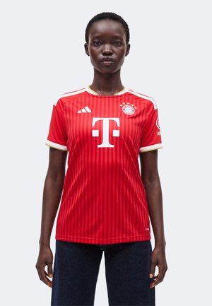 Personne portant un maillot de football rouge du Bayern Munich avec des touches blanches et dorées, debout face à l’avant sur un fond uni.