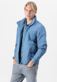 Lys blå jakke med glidelås, høy krage, to sidelommer og ledig passform, kombinert med en matchende polo og grå jeans.