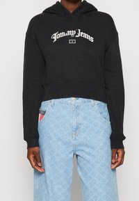 Czarny krótkoterminowy hoodie z kapturem na sznurki i białym logo "Tommy Jeans". W zestawie z jasnoniebieskimi, wzorzystymi dżinsami o luźnym kroju.