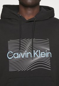 Felpa nera con cappuccio caratterizzata da una grafica frontale con il testo blu "Calvin Klein" e linee ondulate bianche, realizzata in un tessuto morbido con tasca a marsupio.