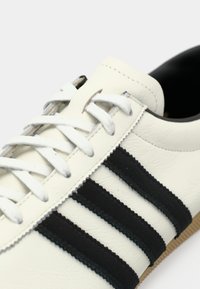 Λευκό δερμάτινο αθλητικό παπούτσι με μαύρες σουέτ λωρίδες, επίπεδα λευκά κορδόνια και υφασμένη λαστιχένια σόλα. Λείο φινίρισμα με στρογγυλή σχεδίαση στη μύτη.