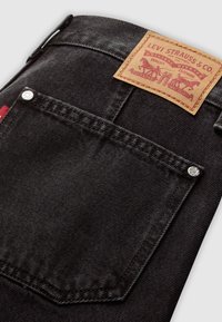 Jeans in denim nero con una tasca posteriore dotata di un rivetto argento e una patch in pelle marrone con la scritta "Levi Strauss & Co" e dettagli grafici.