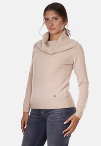 Maglione beige con collo a cowl e maniche lunghe, realizzato in morbido tessuto a maglia. Presenta un sottile logo vicino all'orlo. Indossato con jeans blu scuro.