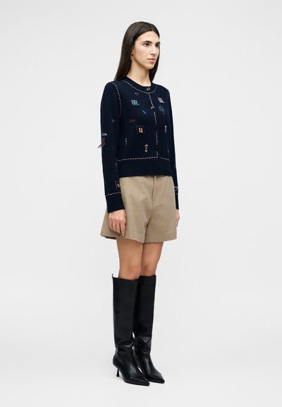 Γυναίκα με μακριά σκούρα μαλλιά που φοράει ένα σκούρο μπλε ζακάρ με κουμπιά και ραμμένα σχέδια, μπεζ σορτς και μαύρες μπότες με τακούνι μέχρι το γόνατο, όρθια.