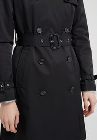 Czarny płaszcz trench z podwójnymi guzikami, z paskiem w talii i błyszczącą klamrą, oraz bocznymi kieszeniami. Gładki materiał o matowym wykończeniu.