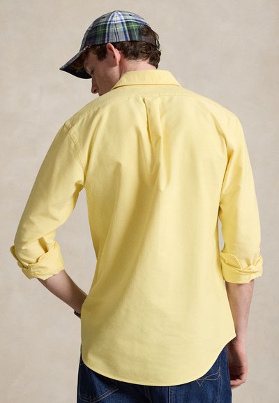 Muž s žlutou košilí s dlouhým rukávem s vyhrnutými rukávy a modrou károvanou čepicí, otočený zády k jednoduchému pozadí.