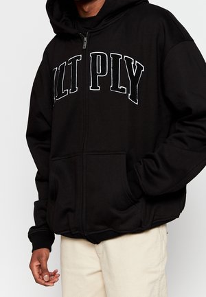 Person trägt einen schwarzen Zip-Hoodie mit weißem "CULT PLY"-Schriftzug, Hände in den Taschen, kombiniert mit hellbeigen Hosen.