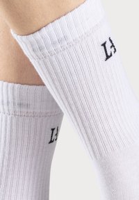 Calcetines blancos acanalados con letras negras en el puño. El diseño es suave con una textura elástica, ideal para la comodidad y el ajuste.