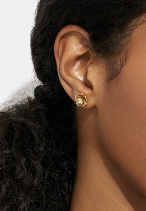Orecchini a forma di fiore in oro, con una gemma trasparente al centro e petali texturizzati che creano un design dettagliato, indossati all'orecchio.
