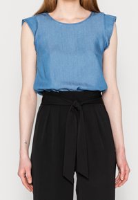 Donna che indossa una blusa blu senza maniche infilata in pantaloni neri a vita alta con una cintura annodata in vita, mostrata dalla nuca ai fianchi.