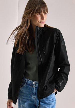 Femme aux longs cheveux portant une veste noire zippée sur un haut sombre et un jean bleu, regardant sur le côté avec une main dans la poche de la veste.