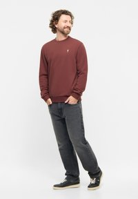 Bordeaux sweatshirt met ronde hals en lange mouwen, voorzien van een klein logo op de borst, gecombineerd met donkergrijze jeans en zwarte sneakers.