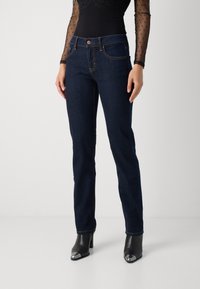 Jeans de mezclilla azul oscuro con silueta ajustada, que cuentan con cinco bolsillos, costuras en contraste y un cierre frontal con botón. Textura suave.