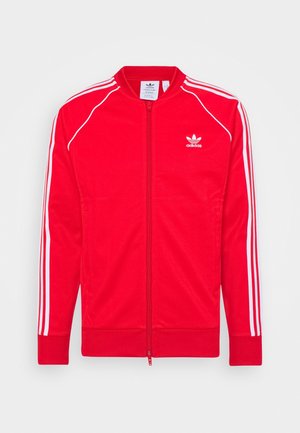 Rode Adidas trainingsjas met witte strepen op de mouwen, een volledige ritssluiting aan de voorkant en het Adidas trefoil-logo op de linkerborst.