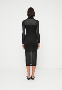 Γυναίκα που στέκεται με την πλάτη προς την κάμερα φορώντας μαύρο διάφανο midi φόρεμα με μακριά μανίκια και μαύρες γόβες με χαμηλό τακούνι σε λευκό φόντο.