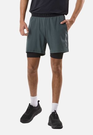 Man wearing black athletic sneakers, white socks, black compression shorts under green running shorts, and a black shirt, standing with hands near pockets.