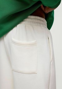 Witte sweatpants met een elastische tailleband en een gestikte achterzak, met een zachte textuur. Groene shirt gedeeltelijk zichtbaar erboven.