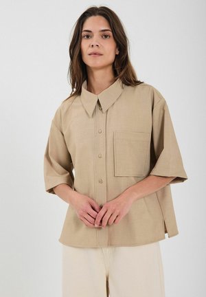 Beige kortärmad skjorta med knäppning fram, spetsig krage, bröstficka och texturerad tyg. Modellen står mot en ljus bakgrund.