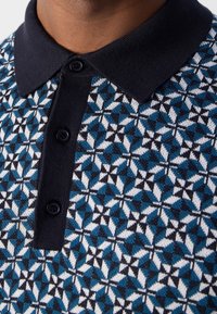 Koszulka polo z granatowym kołnierzykiem i zapięciem na guziki, prezentująca niebiesko-biało-czarny wzór geometryczny. Miękka, teksturowana dzianina.