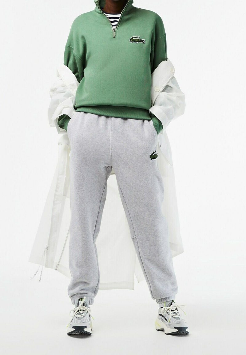 Felpa verde con zip e logo Lacoste, abbinata a pantaloni della tuta grigi con logo. Il modello indossa un cappotto leggero bianco e sneaker.
