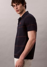 Námořnická polo tričko s červenými horizontálními pruhy, krátkými rukávy a límečkem na dva knoflíky; detail loga na hrudi. Texturovaná bavlněná tkanina.