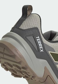 Κοντινό πλάνο της φτέρνας αθλητικού παπουτσιού μονοπατιού σε γκρι και μπεζ χρώμα, με υφή στη σόλα, μαύρη λαβή και τη λέξη "TERREX" στο πλευρικό πάνελ.