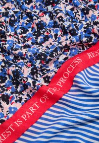 Wielokolorowy materiał z niebiesko-różowo-czarnym wzorem kwiatowym, z czerwonym pasem w talii, na którym widnieje napis "ODPOCZYNEK JEST CZĘŚCIĄ PROCESU" oraz niebieskimi paskami.