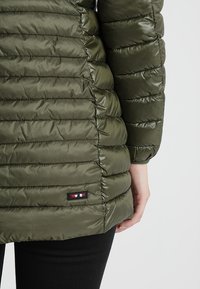 Oliven grøn pufjakke med quiltet tekstur, detalje ved sidesømmen og et lille farvet logo-patch nær kanten. Slim fit, ribbede manchetter synlige.